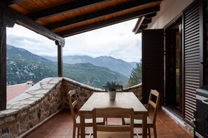 Ein Holztisch mit Stühlen auf einem Balkon, mit einer Steinsäule, mit Blick auf eine gebirgige Landschaft.