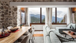 A room with a stone wall, a wooden dining table, a sofa, and a view of mountains through large windows. - Parnassus Woodstone Chalets- N1- N2- N3 (Delphi)