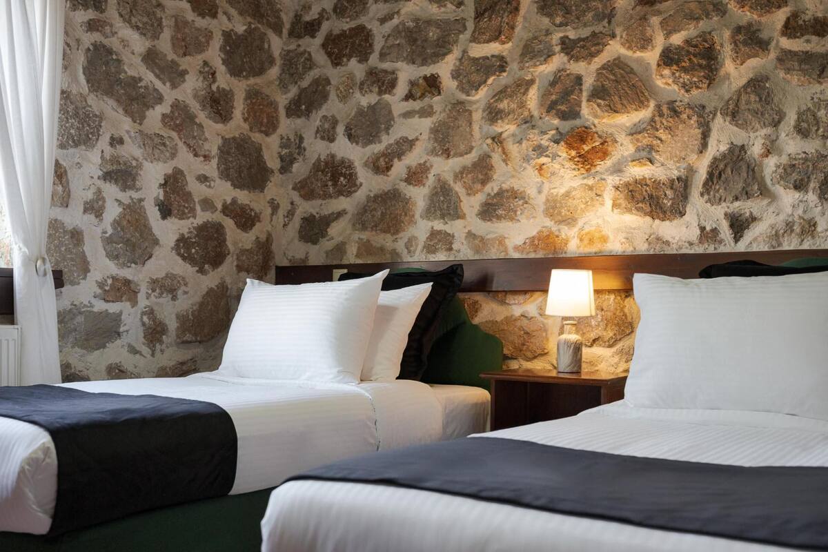Ein Hotelzimmer mit zwei Betten, einer Steinwand, einem Nachttisch mit Lampe und einem Fenster mit Vorhängen.