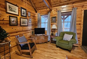 A cozy log cabin interior with wooden walls, a television, a rocking chair, a green armchair, and framed pictures on the wall. - Green Heron in Hocking Hills (Logan)