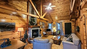 A cozy cabin interior with a fireplace, wooden walls, a ceiling fan, and a television. - Green Heron in Hocking Hills (Logan)