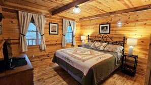 A cozy wooden cabin bedroom with a large bed, two windows with curtains, a bedside table with a lamp, and framed pictures on the walls.