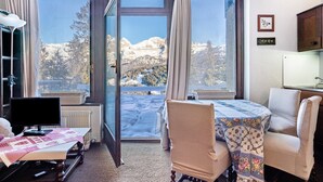 A room with a view of snow-covered mountains, a table with a floral tablecloth, a chair, a desk with a computer, and a radiator.