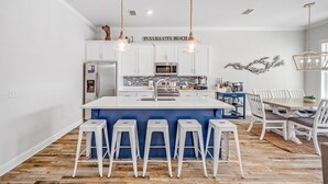 Una cocina moderna con armarios blancos, una isla central con taburetes y un comedor con una isla azul y sillas blancas.