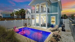 Una casa de dos pisos con piscina, adornada con flamencos rosados, rodeada de mobiliario exterior e iluminada con luces colgantes.