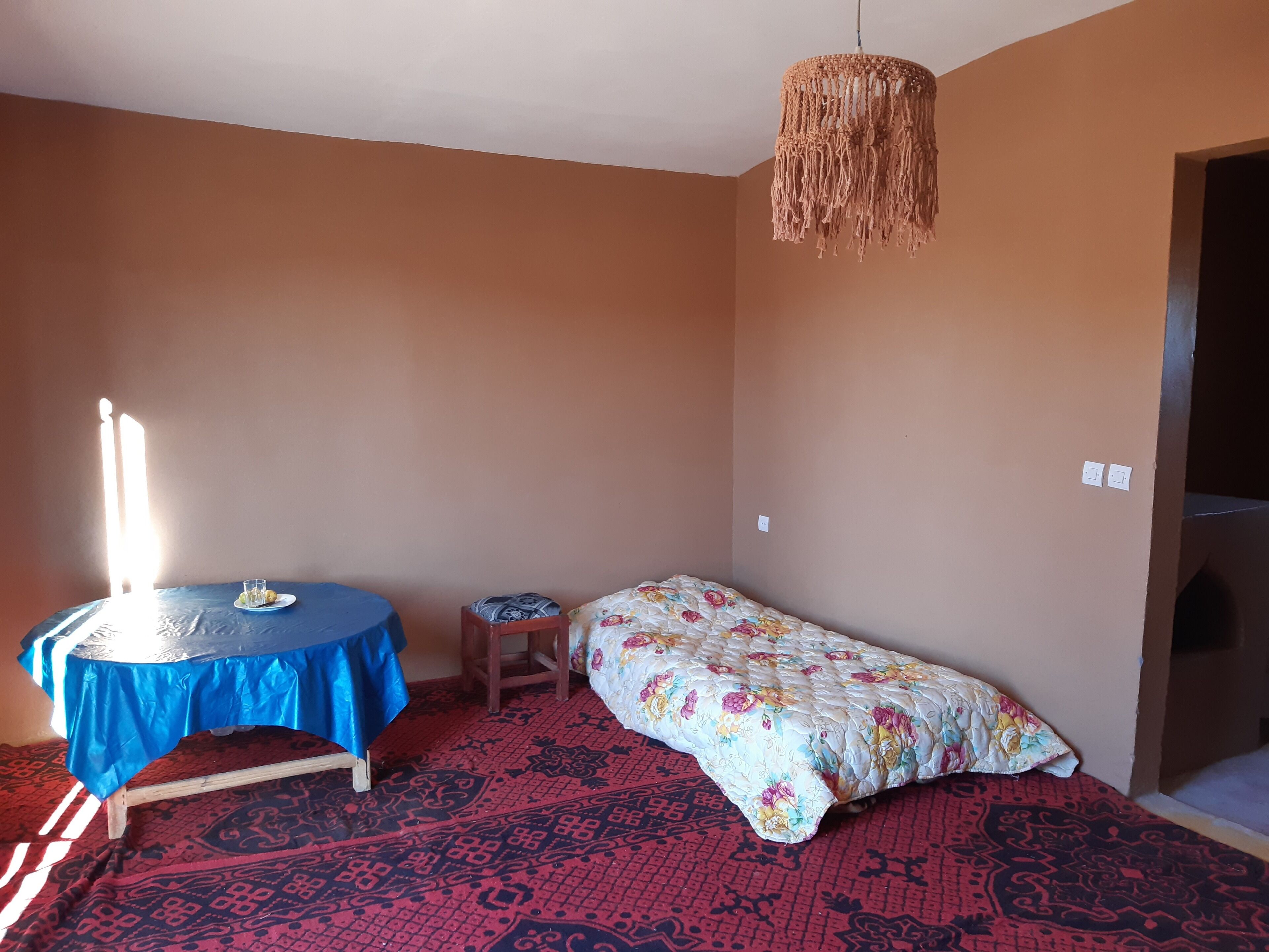 Quarto com cama, uma pequena mesa com toalha de mesa azul, luminária de macramê pendente e carpete estampado.