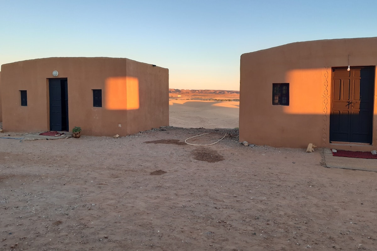 Deux petits bâtiments à un seul étage, dotés de toits plats, sur un sol sablonneux, sous un ciel dégagé.