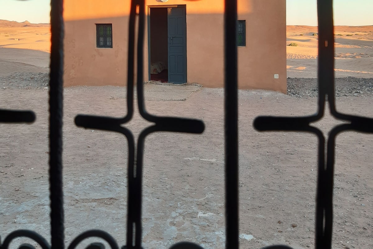 Un petit bâtiment doté d’une porte et d’une fenêtre, vu à travers une grille en métal ornée d’éléments décoratifs.