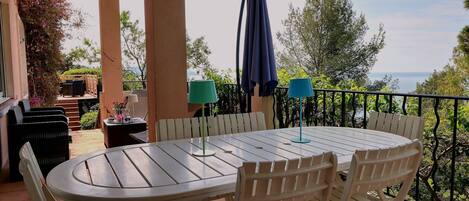 A covered patio with a round table and chairs, a grill, and a view of the sea.