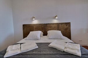 Una cama bien hecha con sábanas y toallas blancas, un cabecero de madera y dos lámparas fijadas a la pared.
