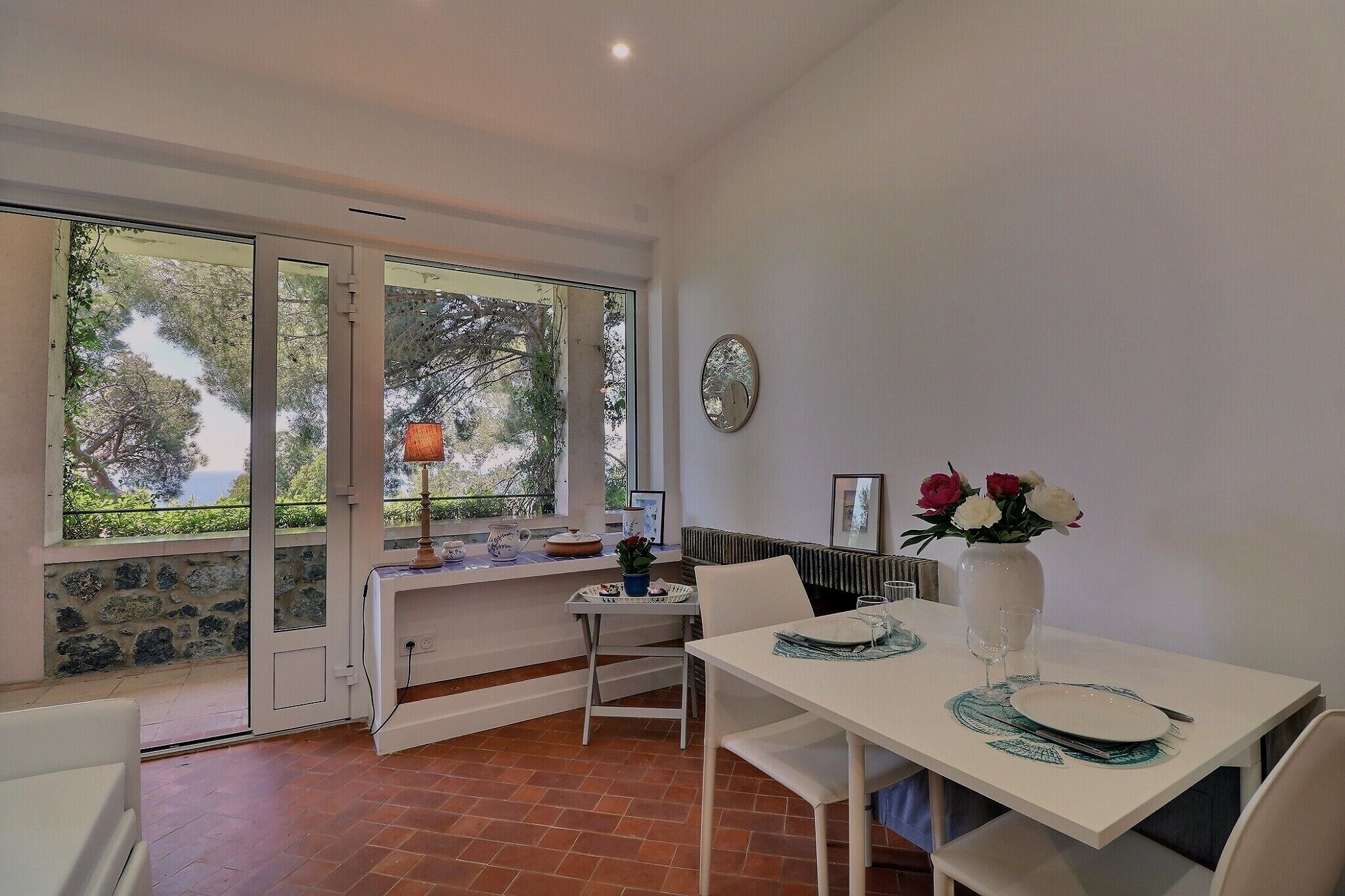 Un coin repas avec une table dressée pour deux personnes, un vase rempli de fleurs et une vue sur les arbres à travers de grandes fenêtres.