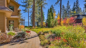 A wooden house with a stone pathway leading to a deck, surrounded by a garden with various plants and flowers.