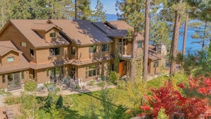 A large, multi-story wooden house with a spacious front yard, surrounded by tall pine trees and a lake view.