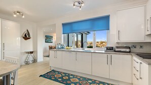 A modern kitchen with white cabinets, a blue window blind, and a view of a coastal town. - Four Oaks - Sea and Harbour Views (Saundersfoot)