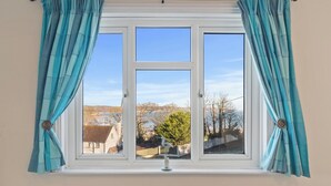 A window with teal curtains, a view of a house, and a bird figurine on the windowsill. - Four Oaks - Sea and Harbour Views (Saundersfoot)