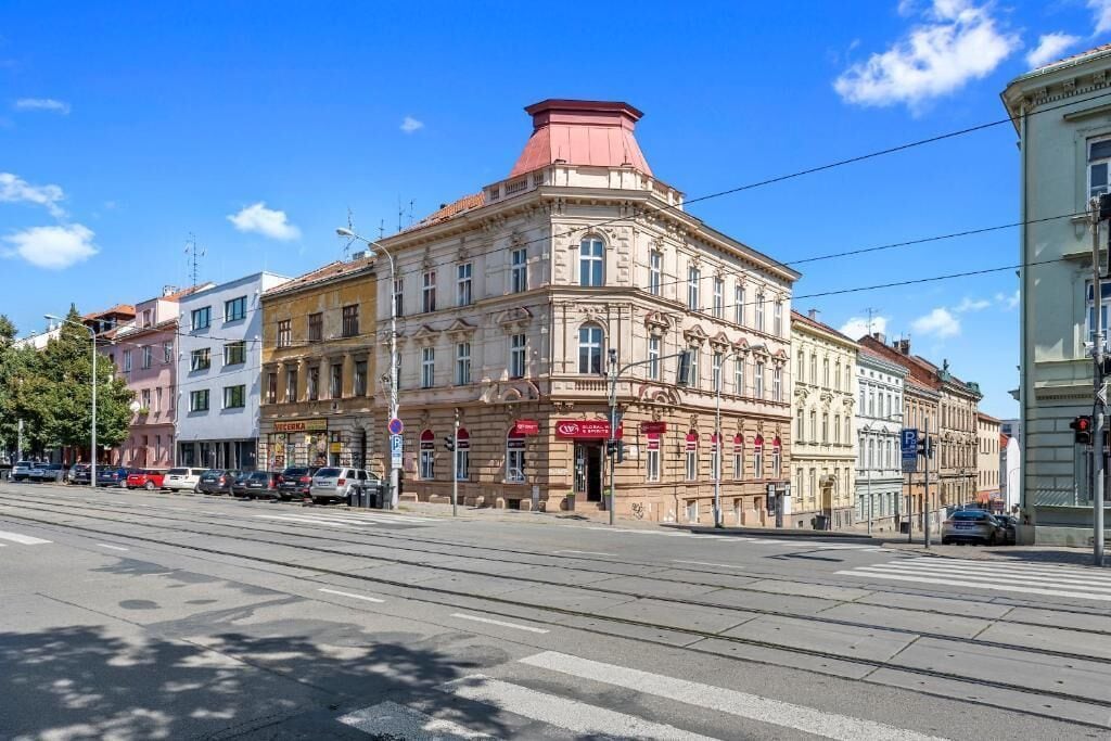 Un edificio histórico con una entrada destacada en esquina, techo rojo y vista a la calle con coches aparcados y una línea de tranvía.