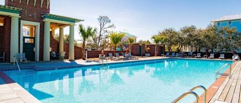 A swimming pool with a wooden deck, lounge chairs, and a brick building with a green roof.