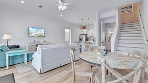 Un salón amplio con un sofá blanco, un sillón azul, una mesa de comedor de madera y una cocina al fondo.