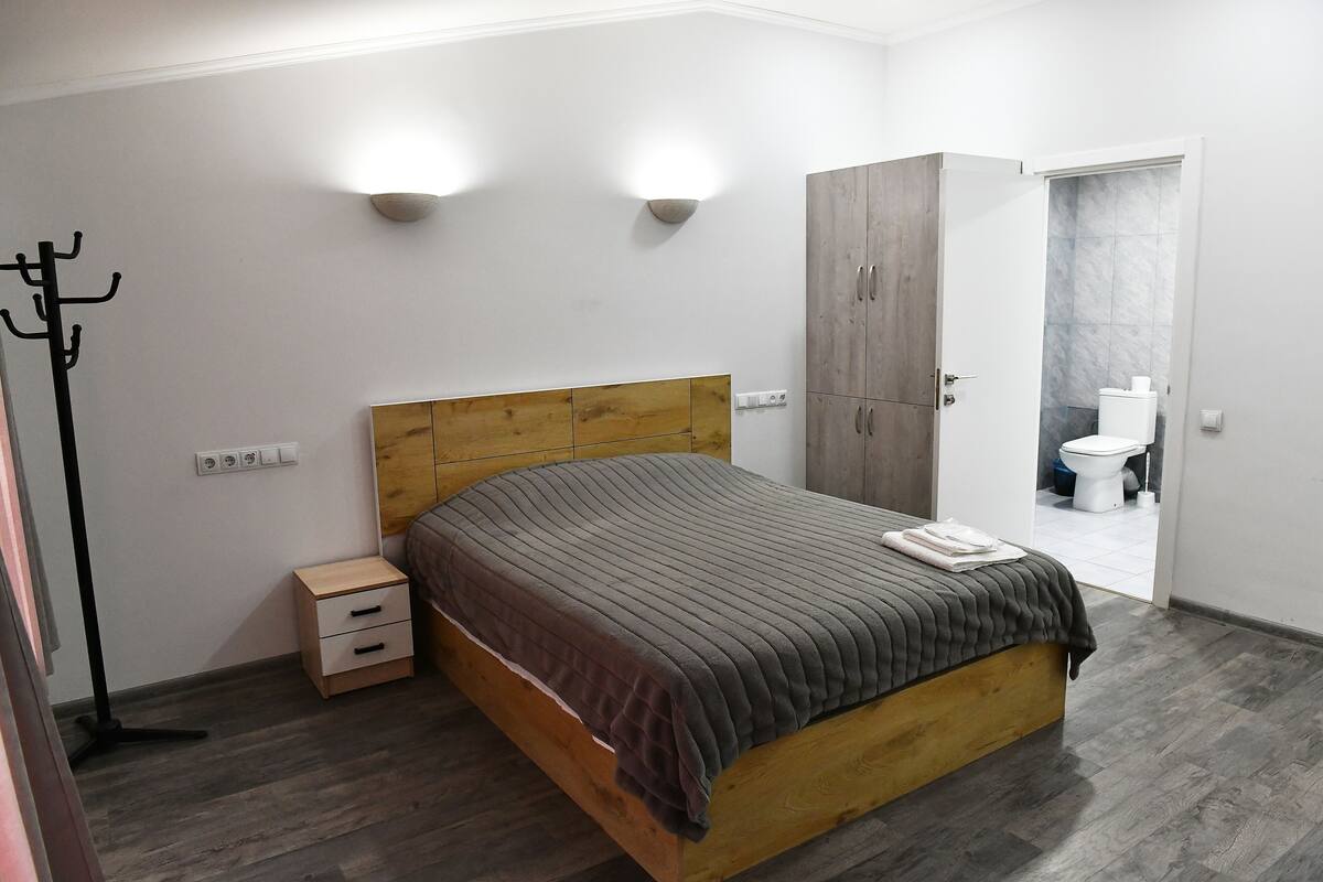 Quarto com cama de madeira, mesas de cabeceira, um cabideiro e um banheiro visível através de uma porta aberta.