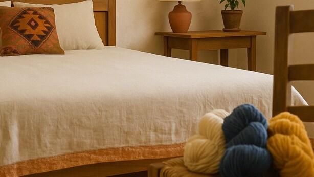 Un letto rifatto con copriletto bianco e cuscino decorato, un comodino in legno con una lampada e una pianta in vaso, e una sedia con gomitoli di lana.