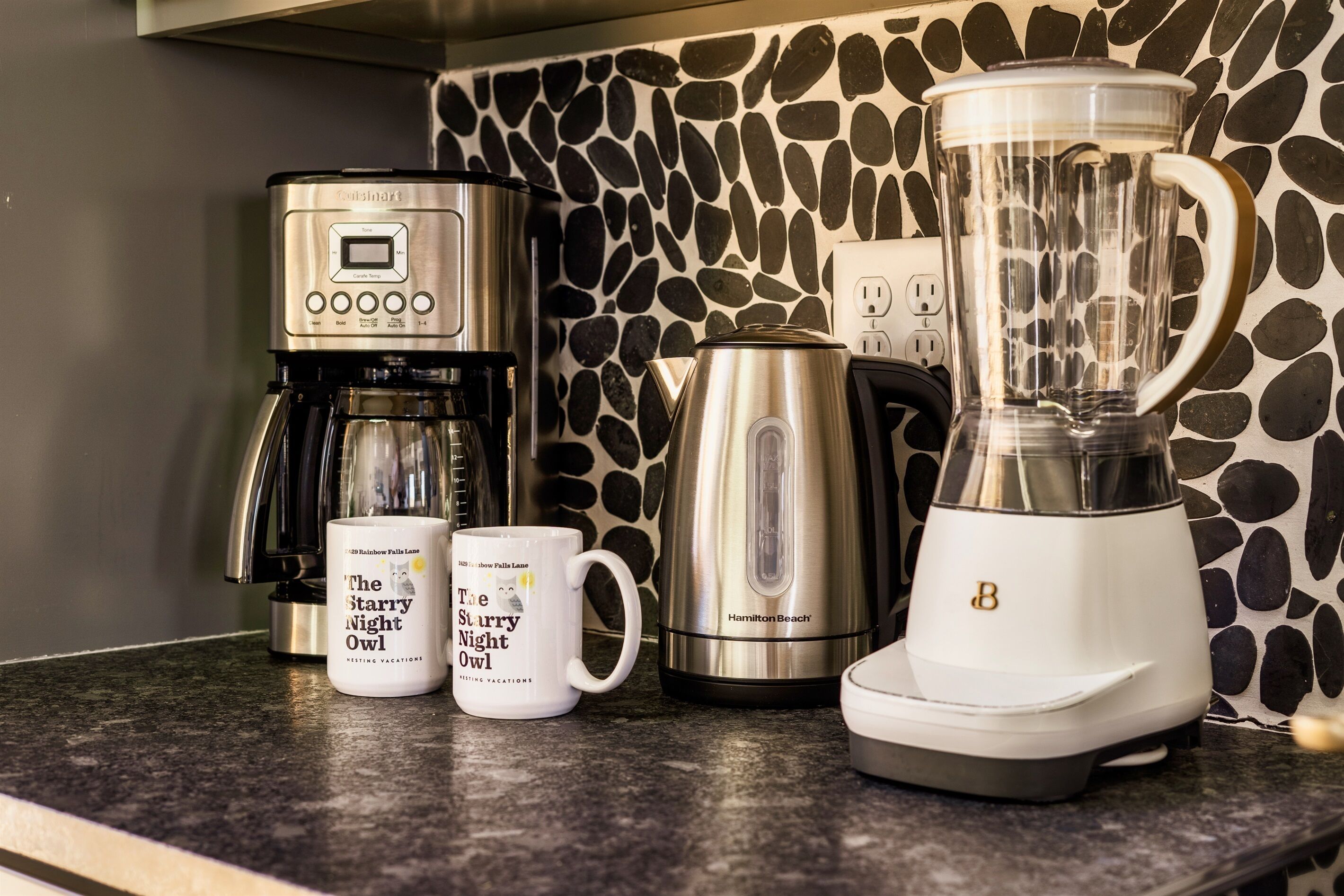 A kitchen counter with a coffee maker, a kettle, a blender, and two mugs.