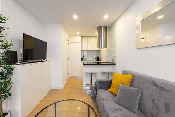 A compact living space with a kitchenette, a grey sofa with yellow and grey cushions, and a flat-screen TV on a white cabinet.