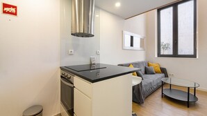 Cocina compacta con estufa, lavamanos y campana extractora de acero inoxidable. Hay un sofá gris con cojines amarillos y una pequeña mesa redonda. Se ve una ventana con una planta.