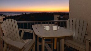 A balcony with two Adirondack chairs and a small table with two white mugs, overlooking a sunset over the ocean. - Oceanfront 3BR,Game Room, Crescent Beach Retreat! (Saint Augustine)