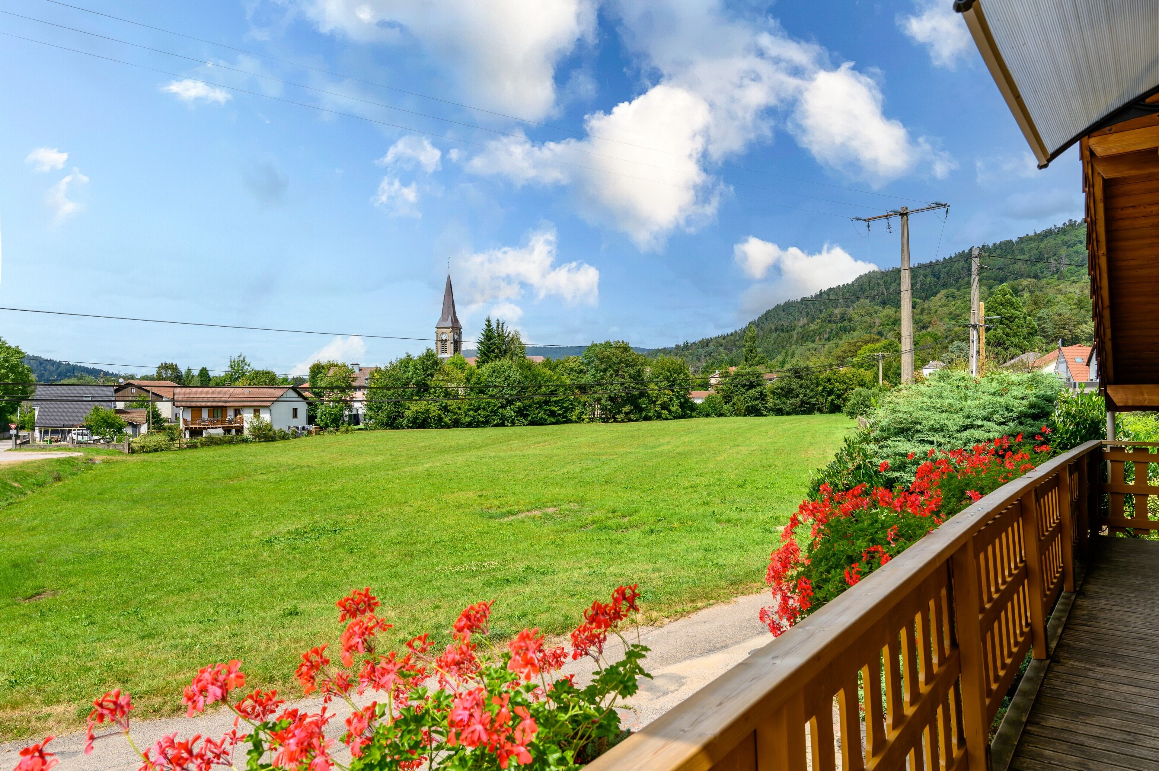 Un champ verdoyant avec une terrasse en bois et des fleurs rouges au premier plan, une église au loin et des maisons sur la gauche.