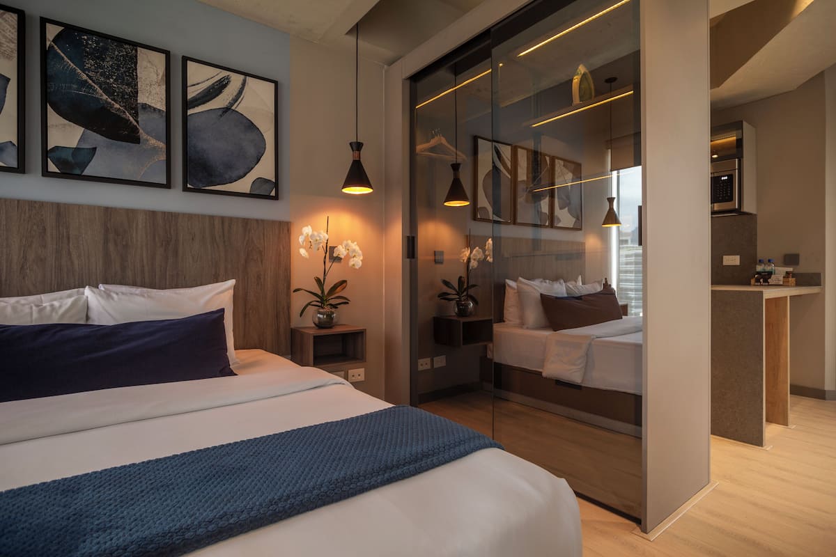 A modern hotel room with a large bed, a separate sleeping area, a kitchenette, and artwork on the walls.