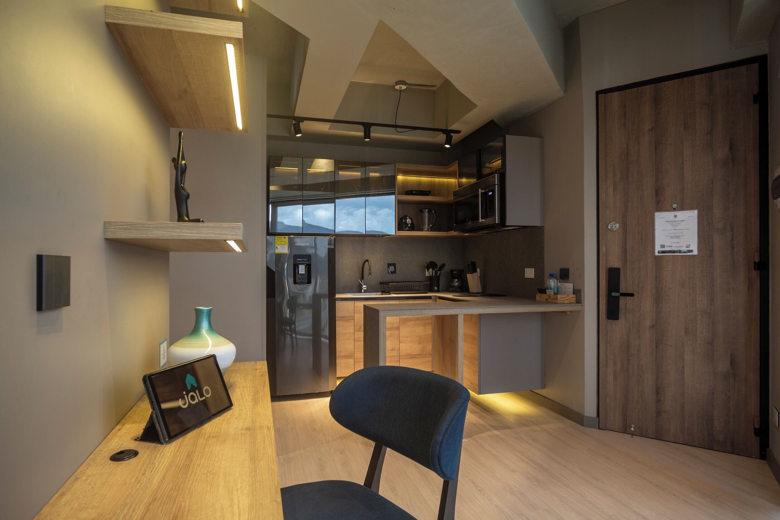 A modern kitchen with a built-in microwave, oven, and sink. There is a wooden door with a sign on it, and a chair in front of the kitchen.