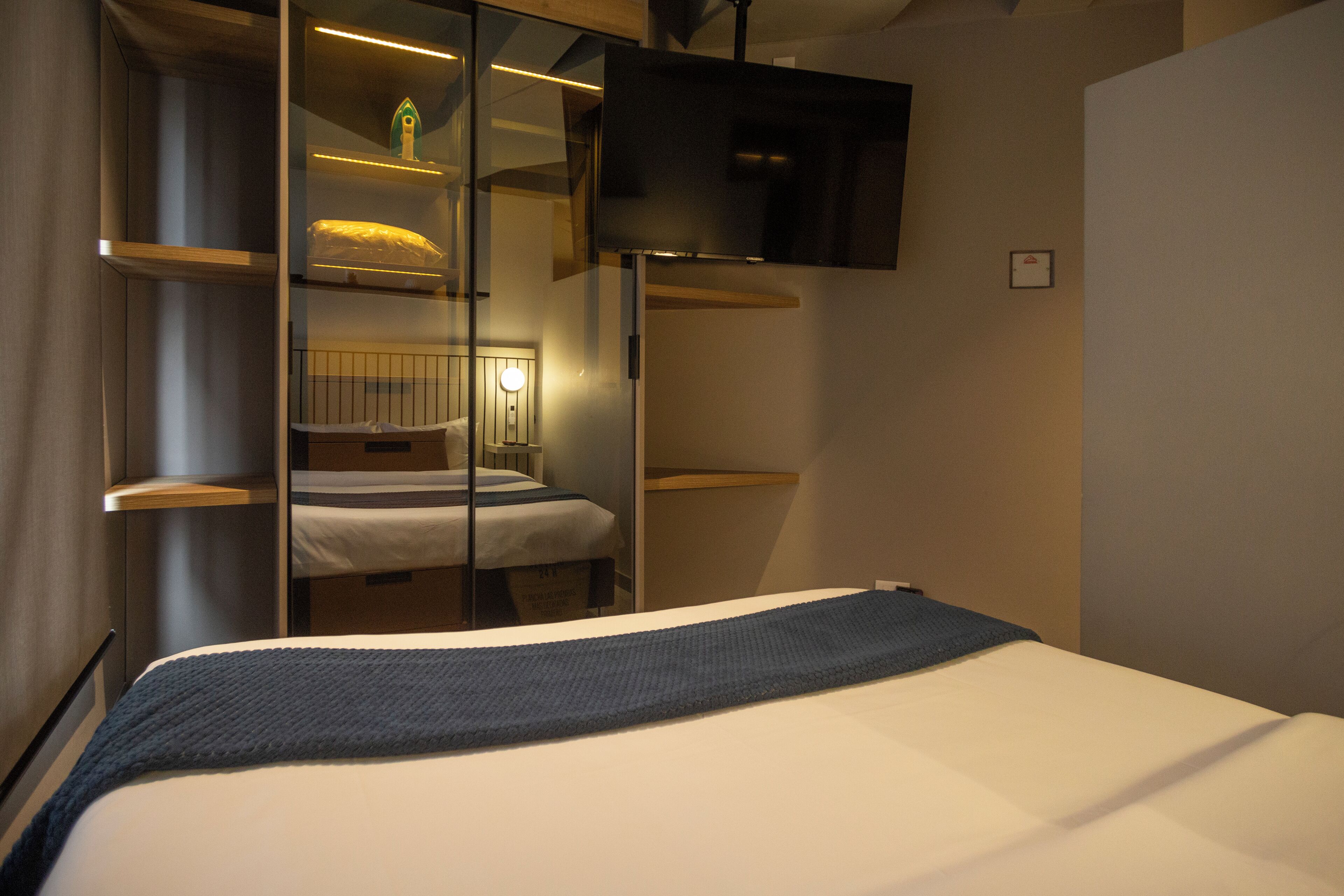 A modern hotel room with a bed, a television mounted on the wall, and a mirror reflecting the room's interior.