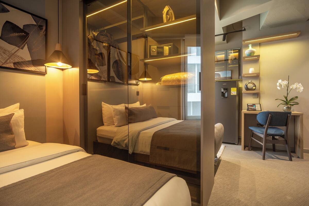 A modern hotel room with a large bed, a desk, and a chair. There is a glass partition between the bed and the desk area.