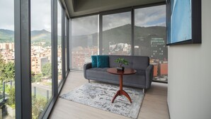Una sala moderna con un sofá gris, una mesa de centro redonda de madera y vistas a un paisaje urbano y montañas a través de ventanas amplias.