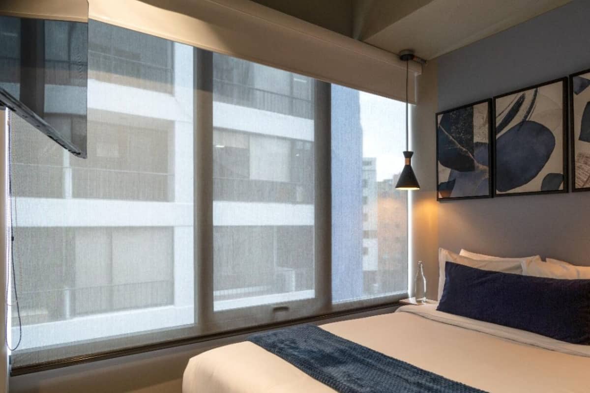 A hotel room with a large window, a bed with white and dark blue bedding, and abstract art on the wall.