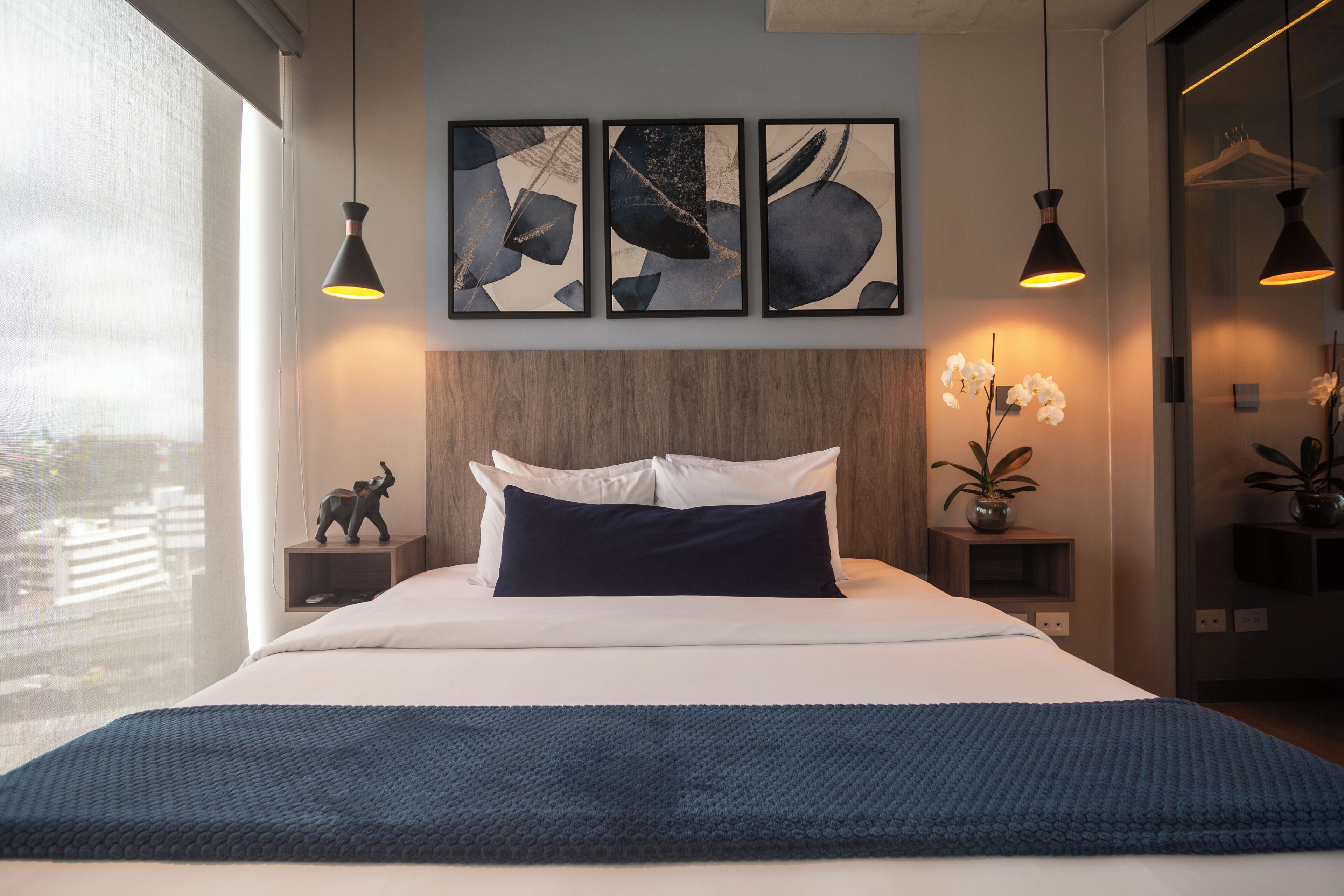 Camera d'albergo moderna con un letto grande, vista sulla città, un'opera d'arte astratta sulla parete e illuminazione a sospensione.