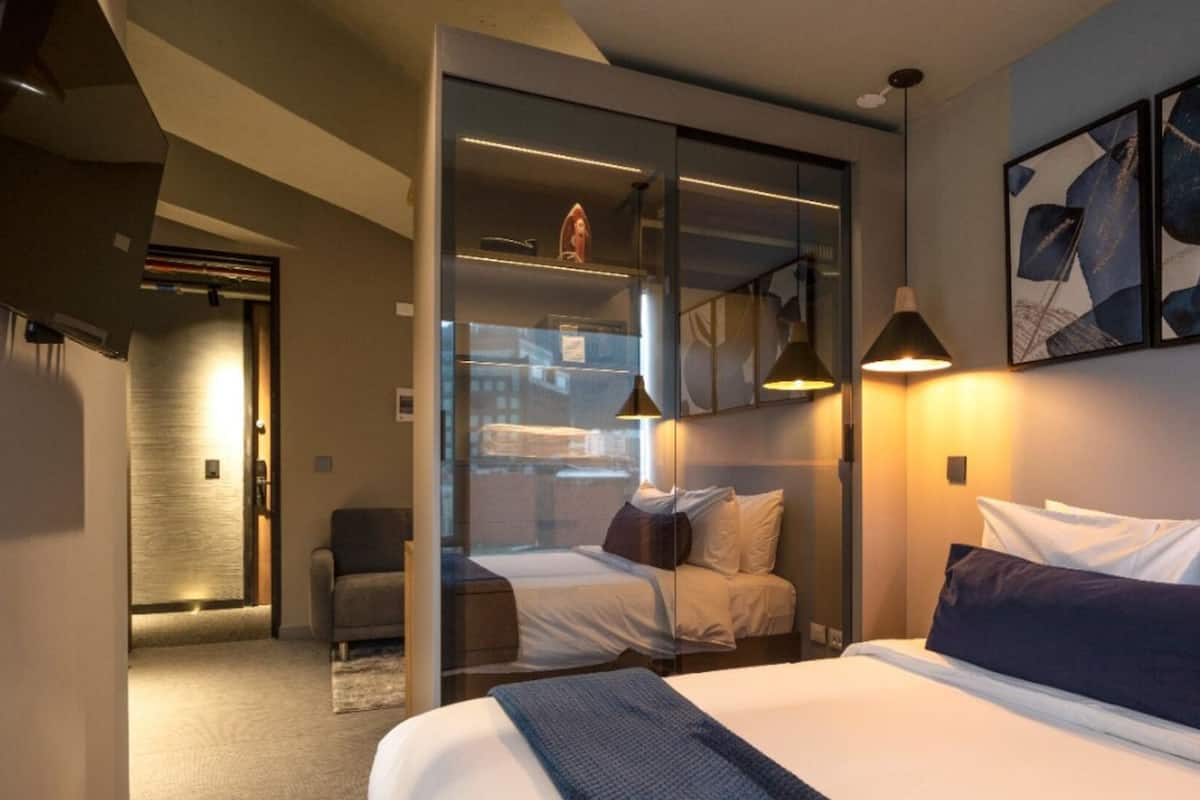 A modern hotel room with a large bed, a seating area, and a glass partition separating the bedroom from the living space.