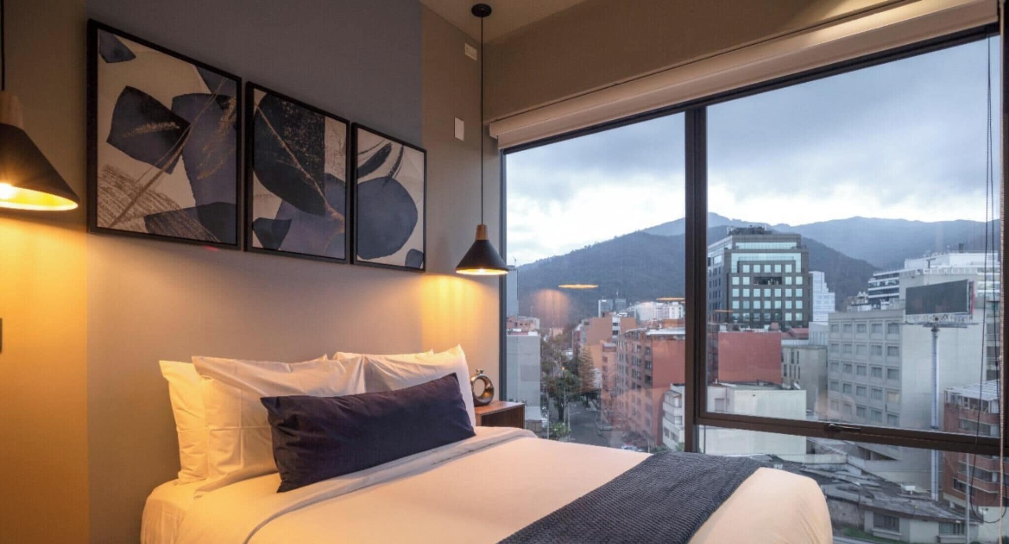 A hotel room with a large bed, two wall-mounted lamps, and a view of a cityscape through a large window.
