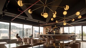 Un bar moderno con techos altos y luminarias geométricas. Varias zonas de estar con mesas y sillas. La barra tiene mostrador y estanterías con botellas. Amplias ventanas ofrecen vistas a la ciudad y a las montañas.