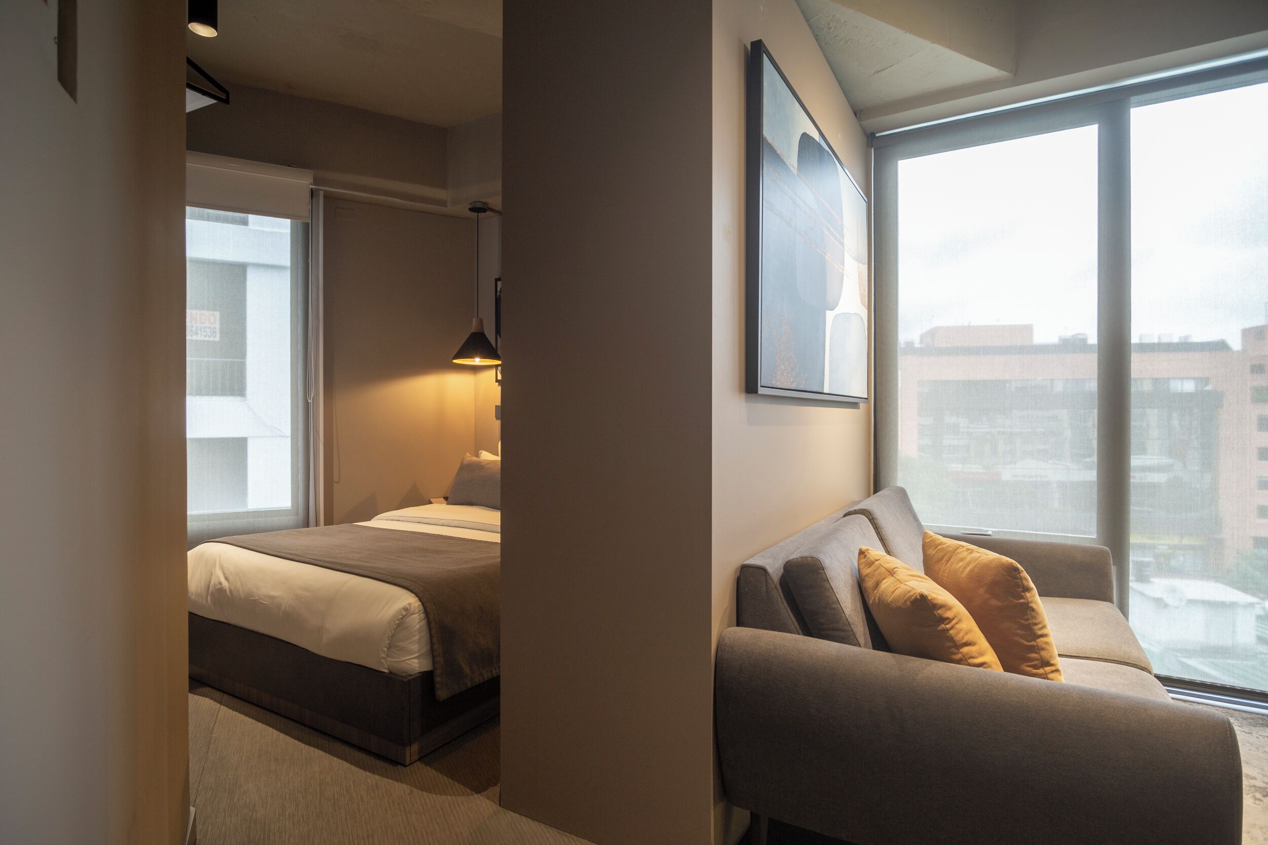 A modern hotel room with a bed, a sofa, and a large window offering a city view.
