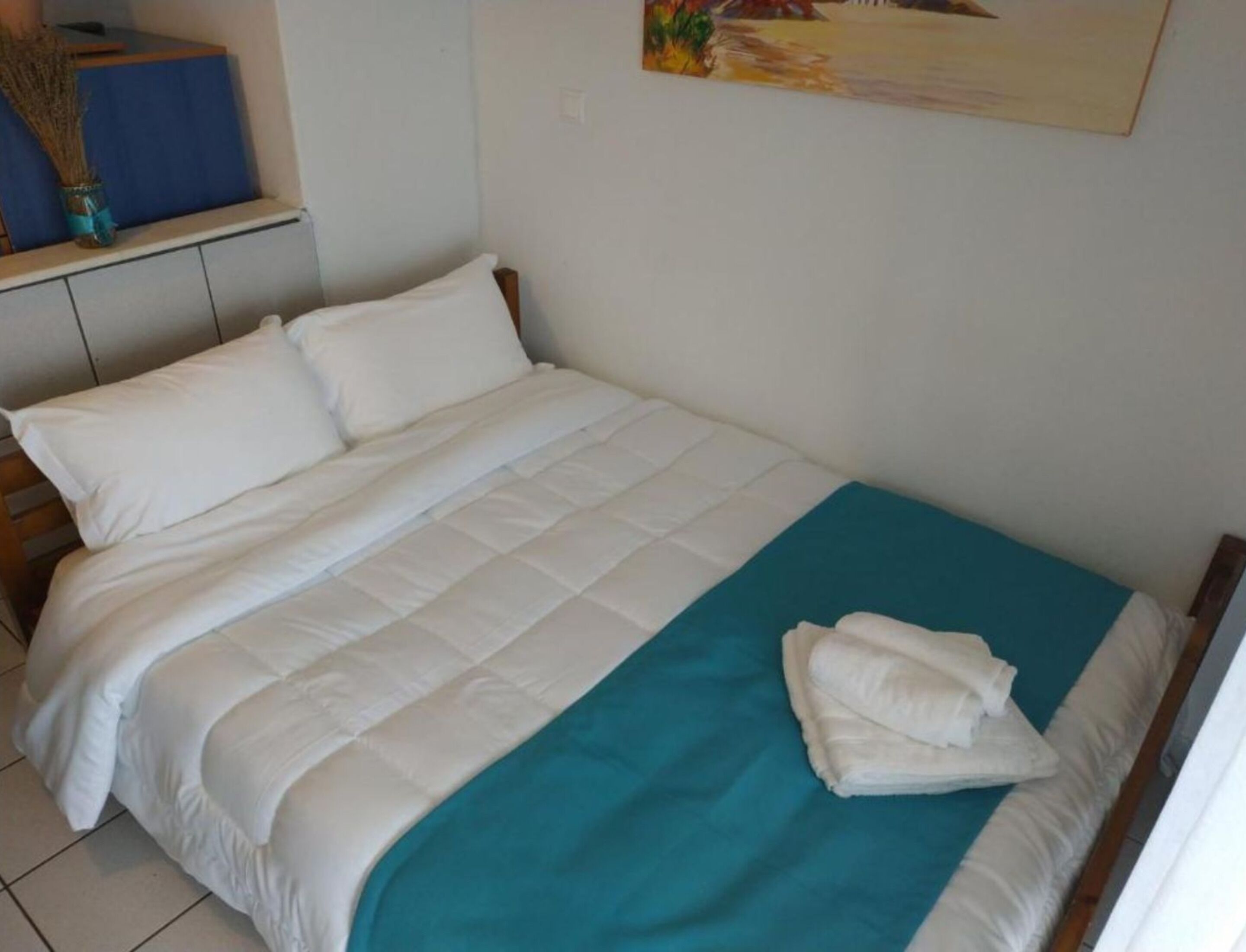 A neatly made bed with white bedding and a teal blanket, a blue wall, and a painting above it.