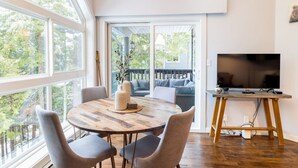A modern living room with a round wooden table, grey chairs, a TV on a wooden stand, and a large window overlooking a deck.