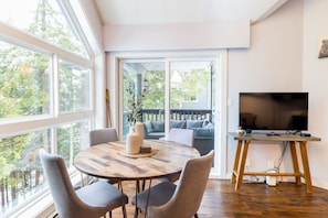 A modern living room with a round wooden table, grey chairs, a TV on a wooden stand, and a large window overlooking a deck.