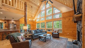 A spacious living area with a high ceiling, wooden walls, a stone fireplace, and large windows offering a view of the outdoors. - Cozy Log Chalet Near Wisp w/ Hot Tub (McHenry)