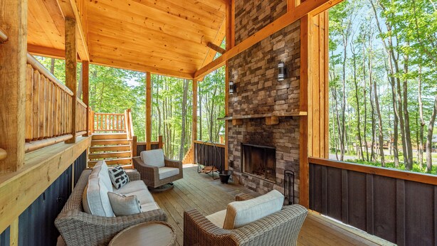A wooden deck with wicker furniture, a stone fireplace, and a view of the forest. - Cozy Log Chalet Near Wisp w/ Hot Tub (McHenry)