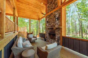 A wooden deck with wicker furniture, a stone fireplace, and a view of the forest. - Cozy Log Chalet Near Wisp w/ Hot Tub (McHenry)