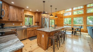 A spacious kitchen with wooden cabinets, stainless steel appliances, a granite countertop, and a large island with bar stools. - Cozy Log Chalet Near Wisp w/ Hot Tub (McHenry)