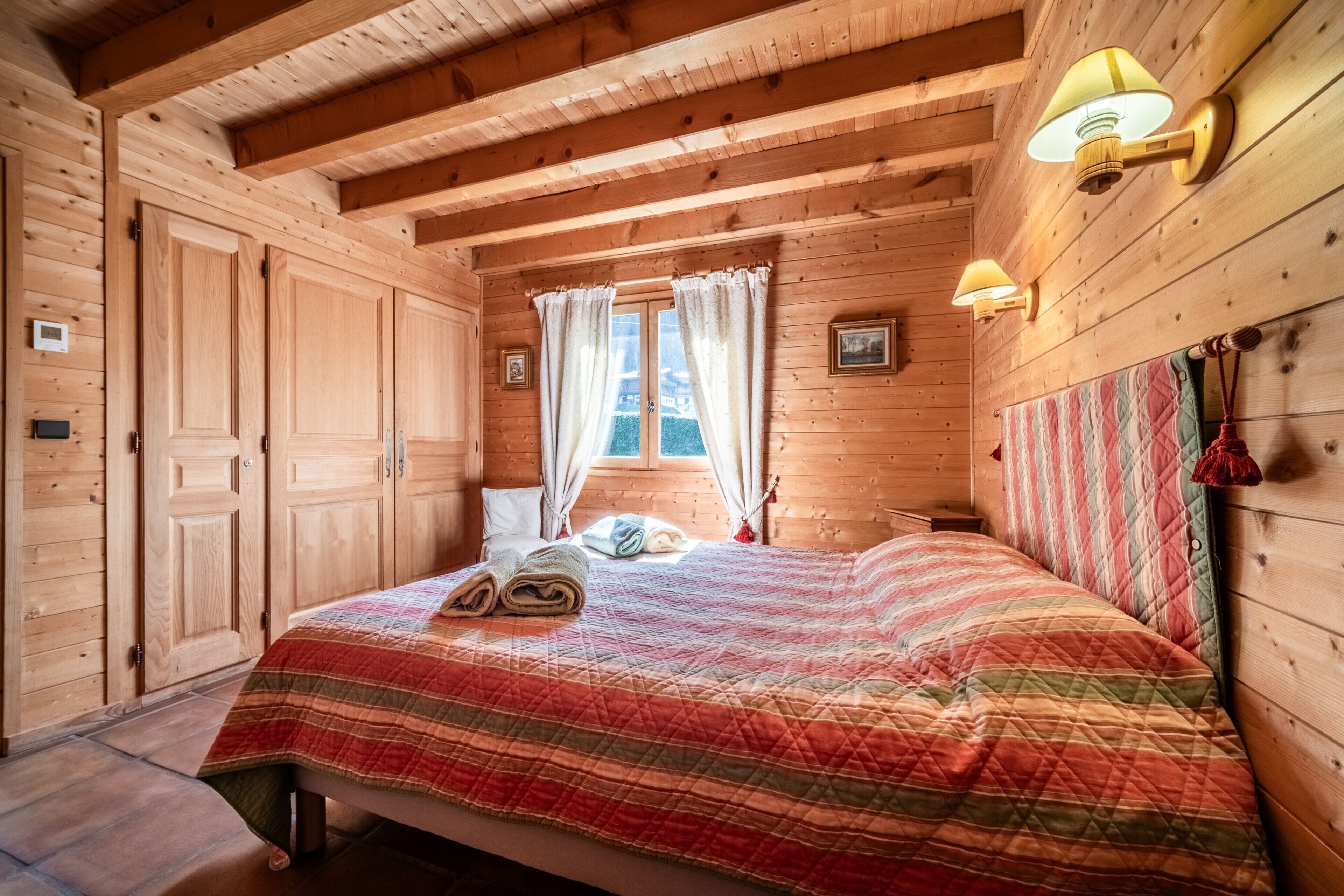 A wooden cabin room with a bed, two wall-mounted lamps, a window with curtains, and a quilted bedspread.