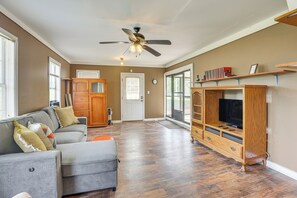 Un salon spacieux doté d’un plancher en bois franc, d’un canapé d’angle gris, d’un meuble de rangement en bois pour les appareils électroniques et d’un ventilateur de plafond.