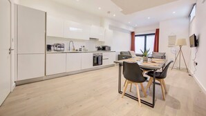 Een moderne keuken met witte kastjes, een eethoek met een tafel en stoelen, en een groot raam met gordijnen.